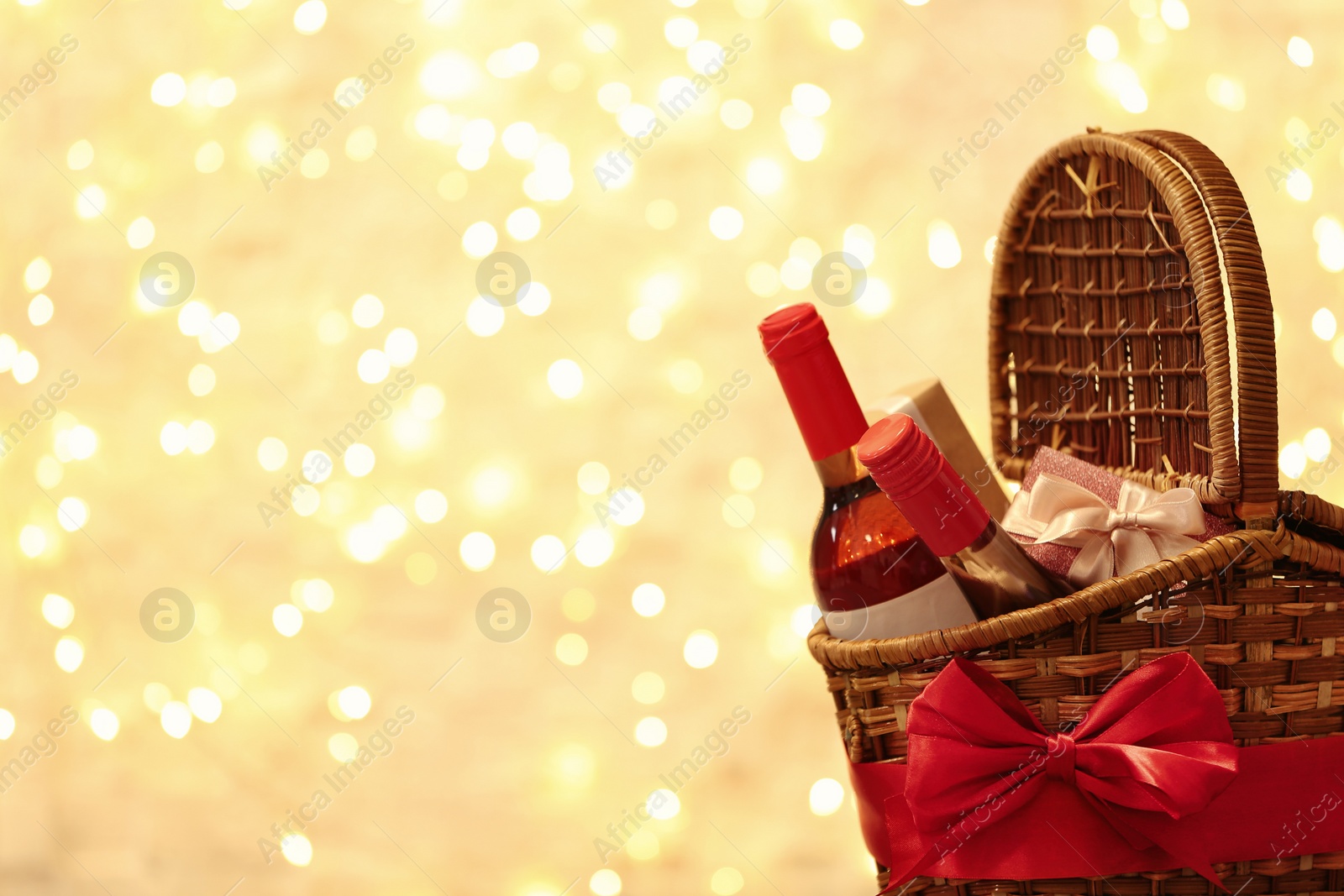 Photo of Festive basket with bottles of wine and gifts against blurred lights. Space for text