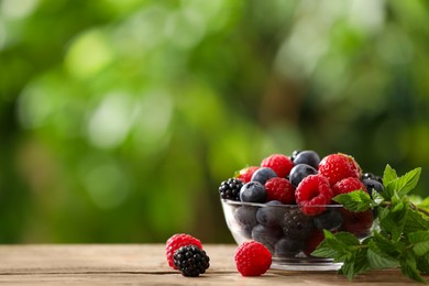 Glass bowl with different fresh ripe berries and mint on wooden table outdoors, space for text