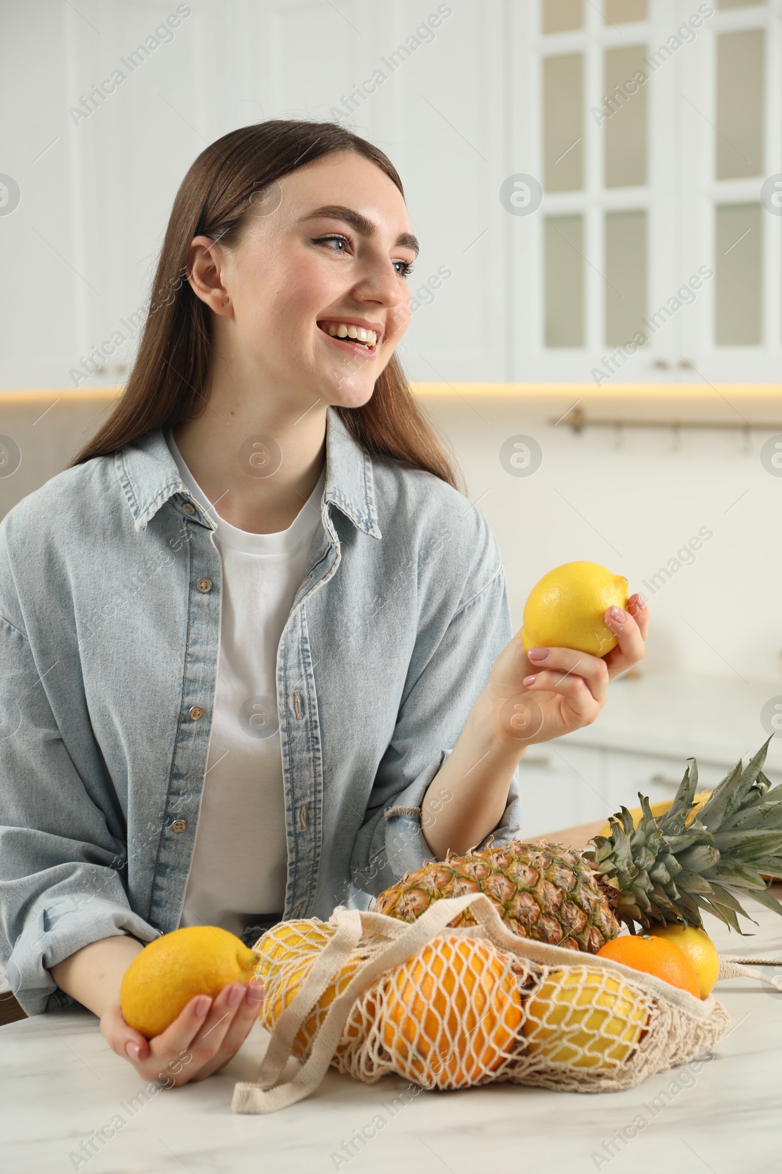 Photo of Woman with lemons and string bag of fresh fruits at light marble table in kitchen