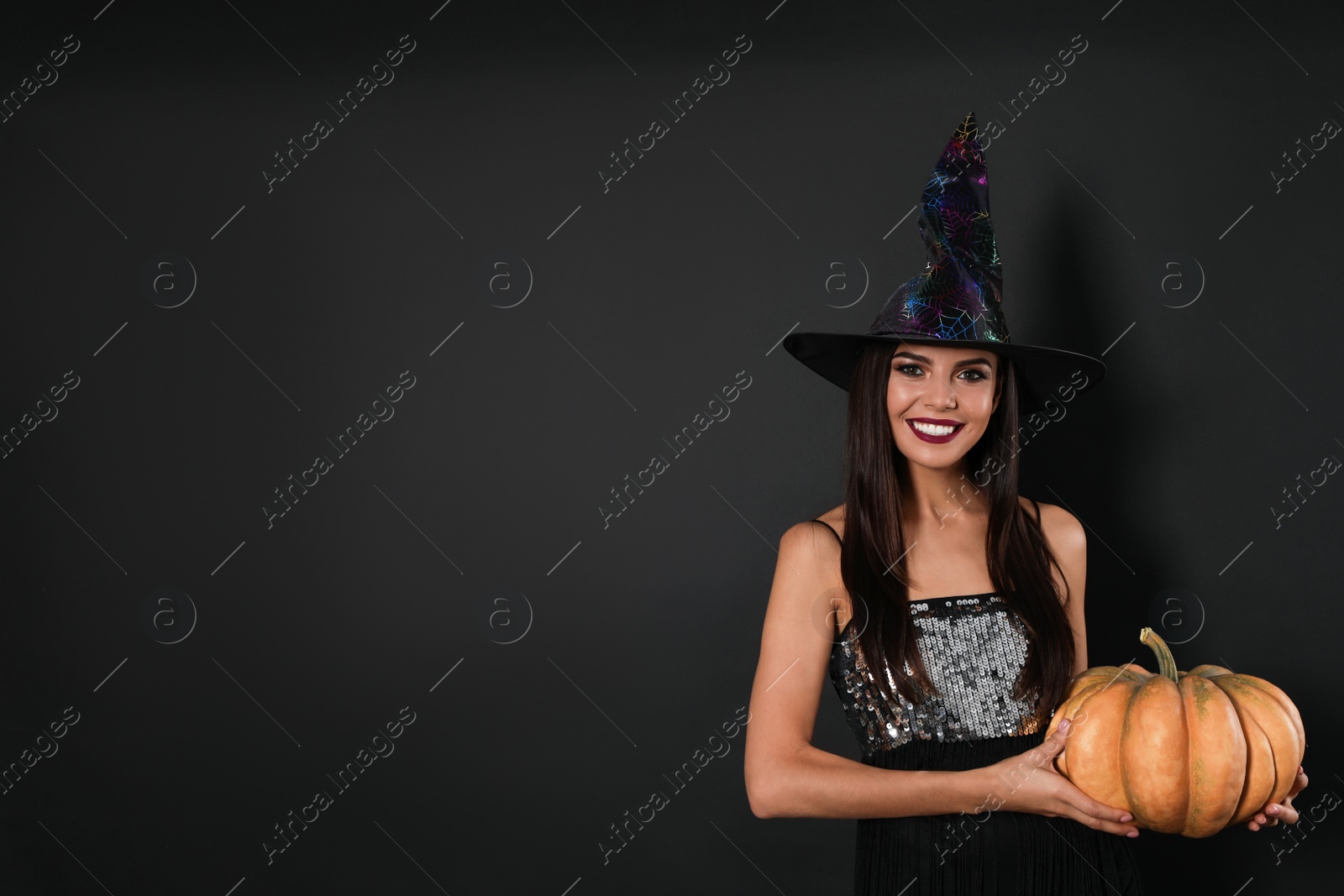 Photo of Beautiful woman wearing witch costume with pumpkin for Halloween party on black background, space for text