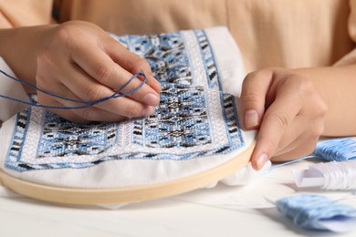 Photo of Woman embroidering shirt with blue thread at table, closeup. Ukrainian national clothes