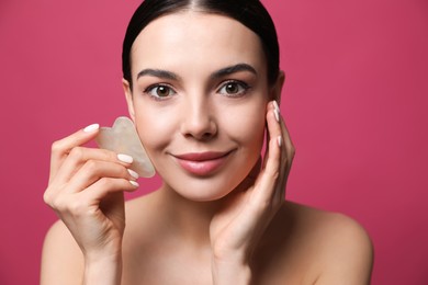 Beautiful young woman doing facial massage with gua sha tool on pink background, closeup