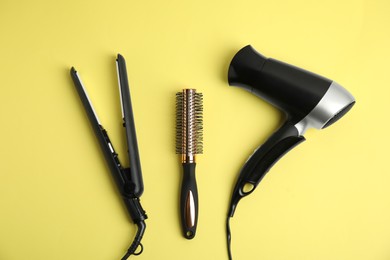 Photo of Hair dryer, straightener and brush on yellow background, flat lay
