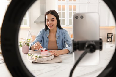 Photo of Blogger with cake recording video on smartphone camera in kitchen at home, view through ring lamp