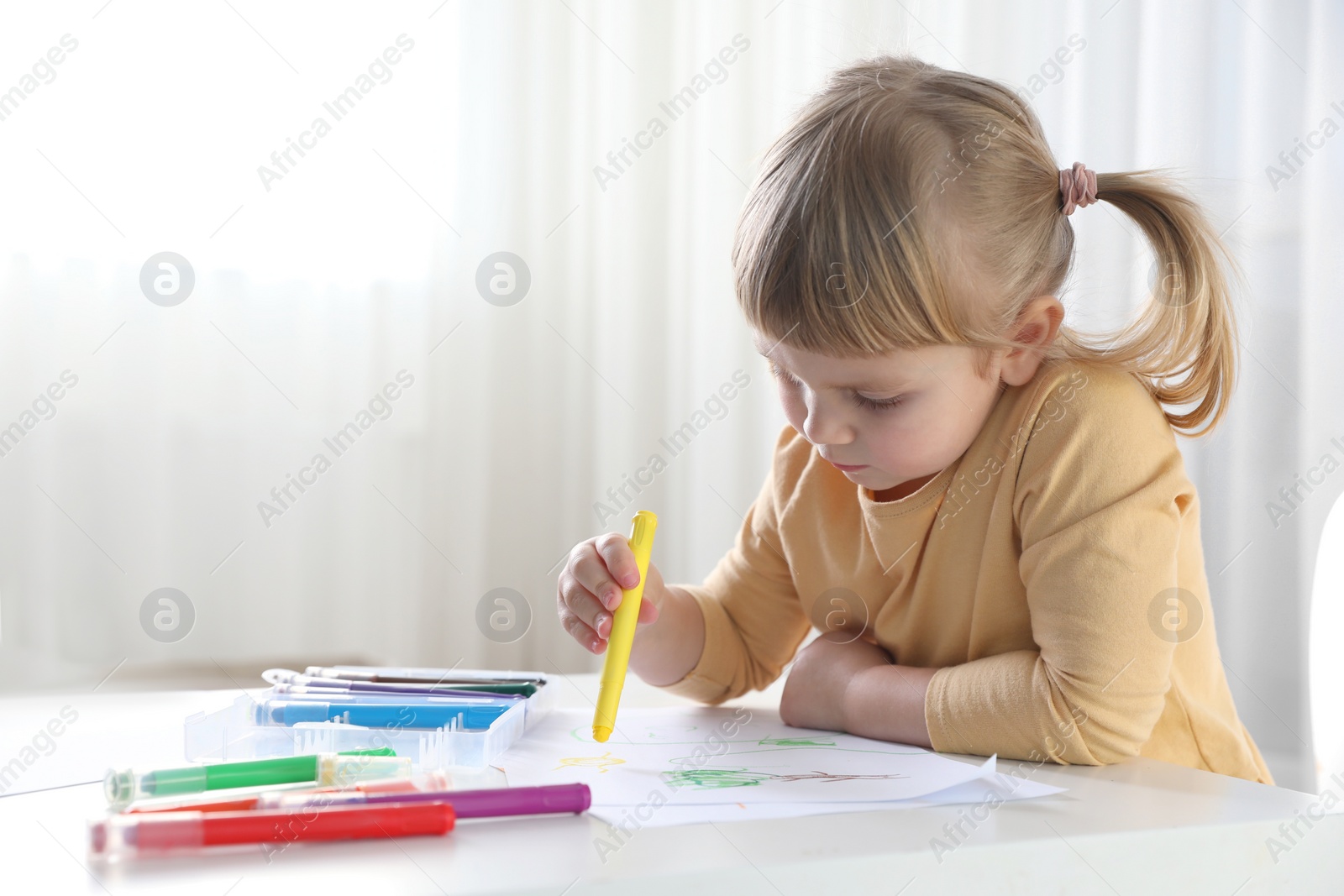Photo of Cute little girl drawing with marker at white table indoors. Child`s art
