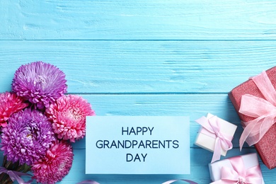 Photo of Beautiful flowers, gift boxes and card with phrase Happy Grandparents Day on light blue wooden background, flat lay. Space for text