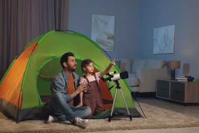 Photo of Father and his daughter using telescope to look at stars while sitting in camping tent indoors