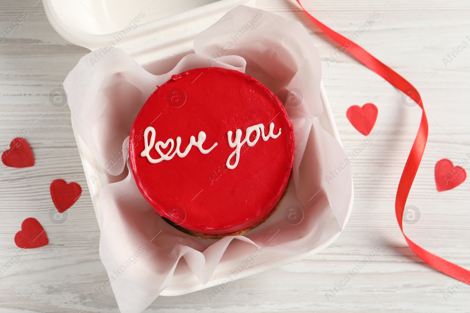 Photo of Bento cake with Love You text in takeaway box and paper hearts on white wooden table, flat lay. St. Valentine's day surprise