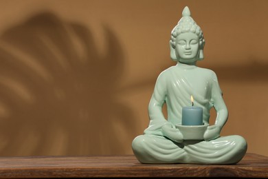 Photo of Buddhism religion. Decorative Buddha statue with burning candle on wooden table against light brown wall, space for text