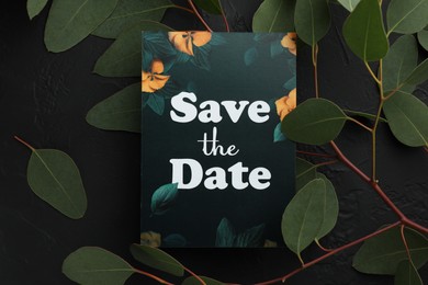 Photo of Beautiful card with Save the Date phrase and green leaves on black textured background, flat lay