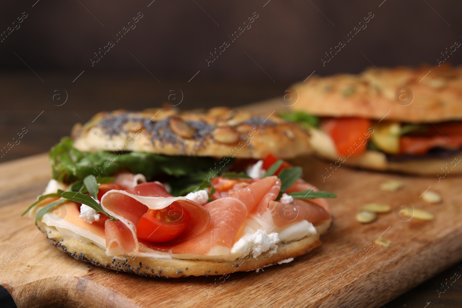 Photo of Tasty bagel with cured ham, cream cheese, tomatoes and arugula on wooden board, closeup