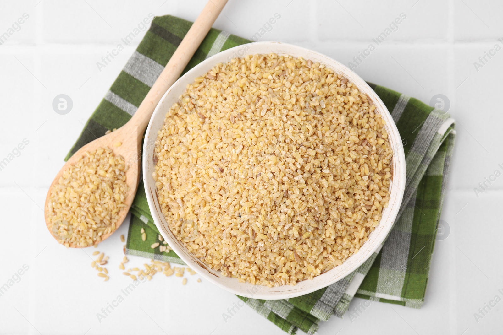 Photo of Bowl and spoon with raw bulgur on white tiled table, top view