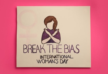 Photo of Card with text Break The Bias, International Women's Day and painting of girl showing crossed arms on pink background, top view