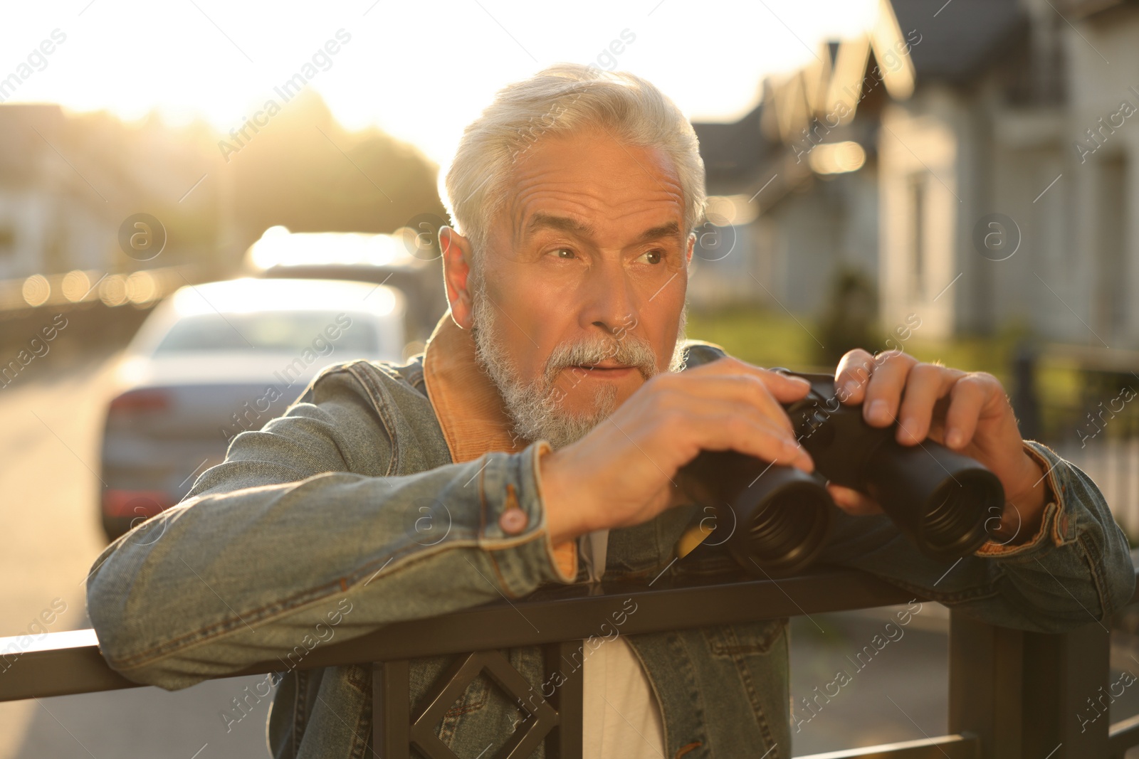 Photo of Concept of private life. Curious senior man with binoculars spying on neighbours over fence outdoors