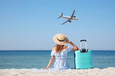 Woman with suitcase on beach looking at airplane flying in sky, back view