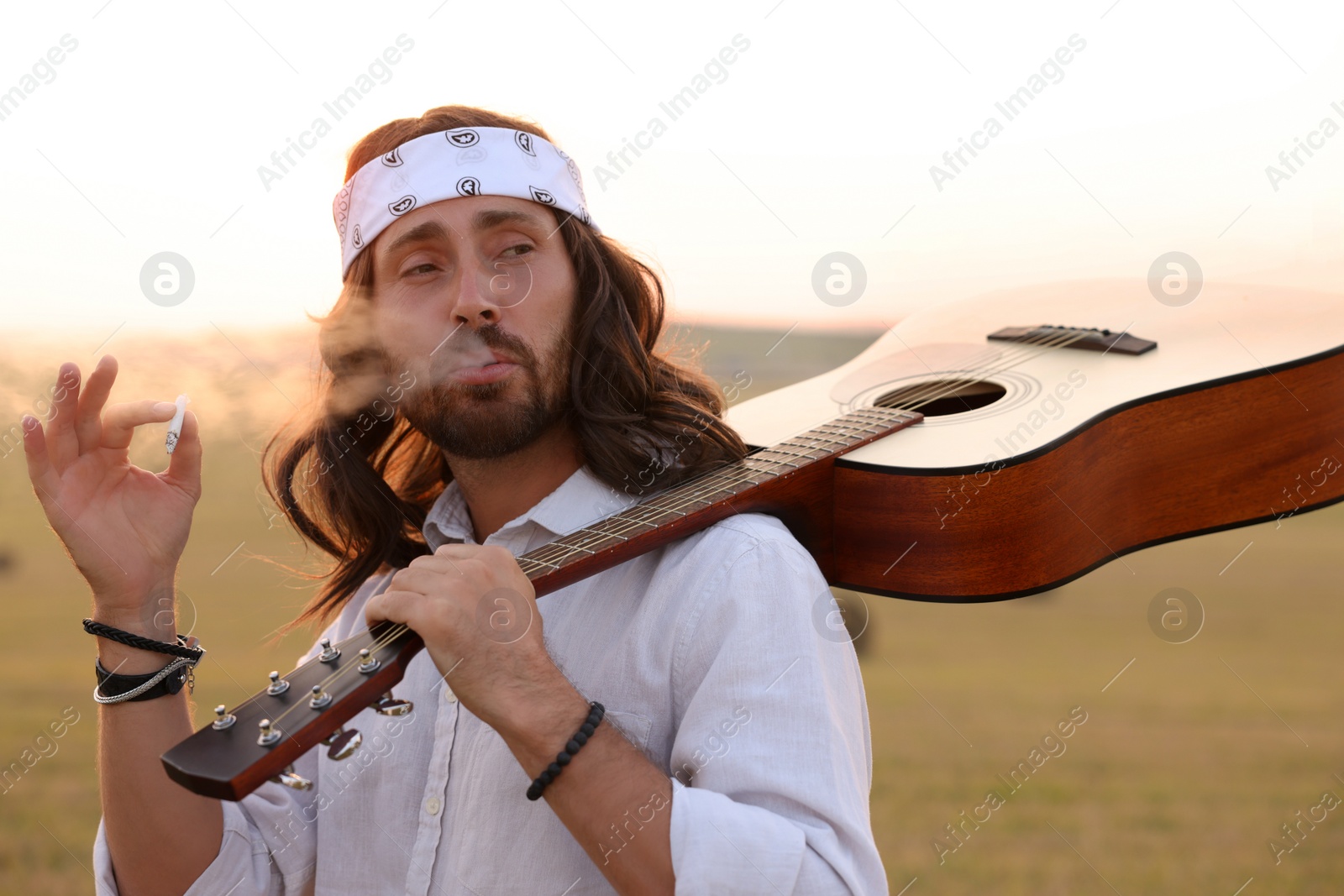 Photo of Stylish hippie man with guitar smoking joint in field