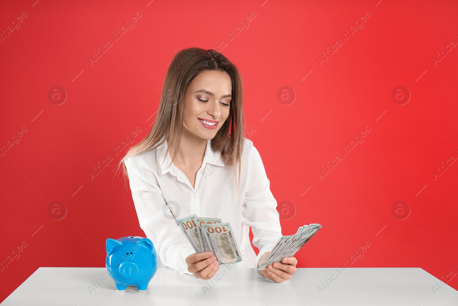 Photo of Young woman with money and piggy bank at table on crimson background