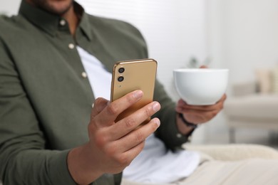 Photo of Man with cup of drink sending message via smartphone indoors, closeup