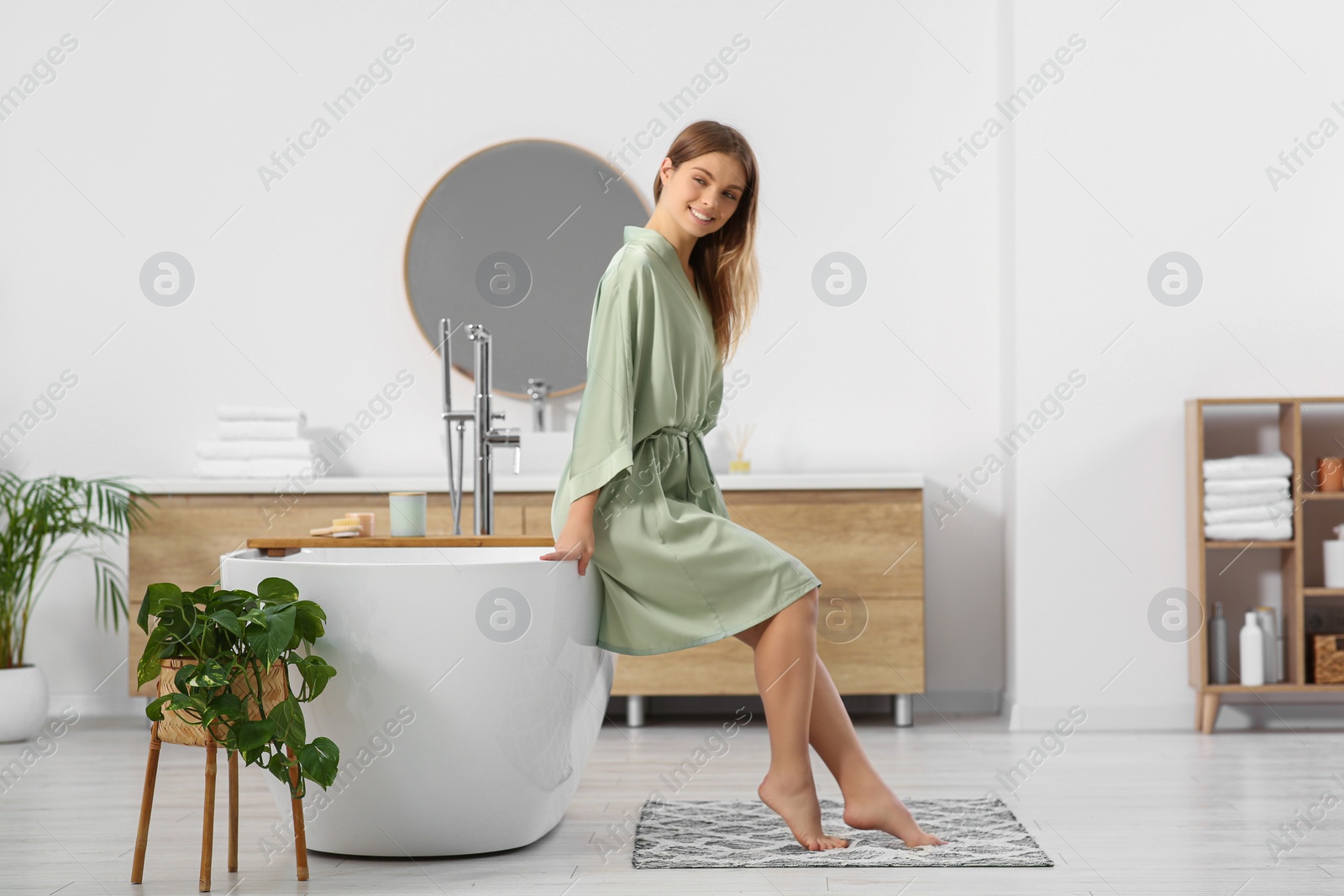 Photo of Beautiful happy woman in stylish robe sitting on tub in bathroom