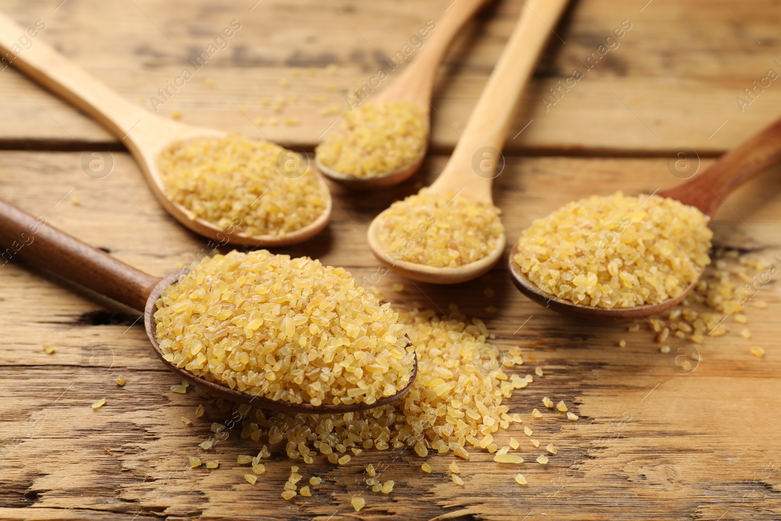 Photo of Spoons with raw bulgur on wooden table, closeup