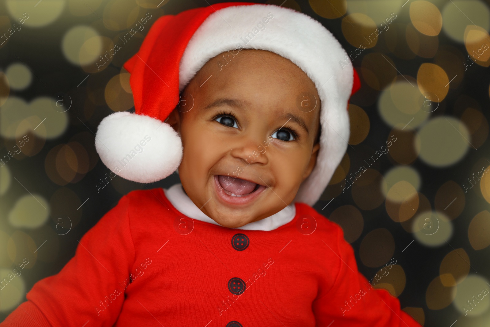 Image of Cute little African American baby wearing Santa hat against blurred lights on dark background. Christmas celebration