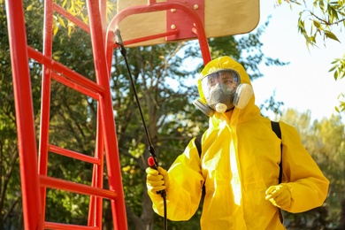 Photo of Person in hazmat suit with disinfectant sprayer cleaning children's playground. Surface treatment during coronavirus pandemic