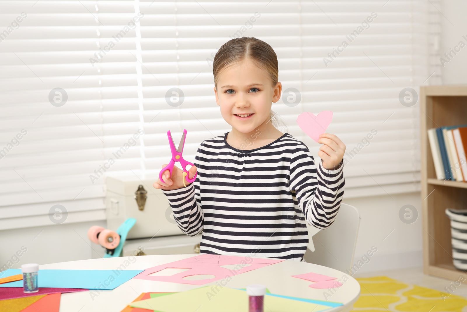 Photo of Cute little girl with pink paper heart and scissors at desk in room. Home workplace
