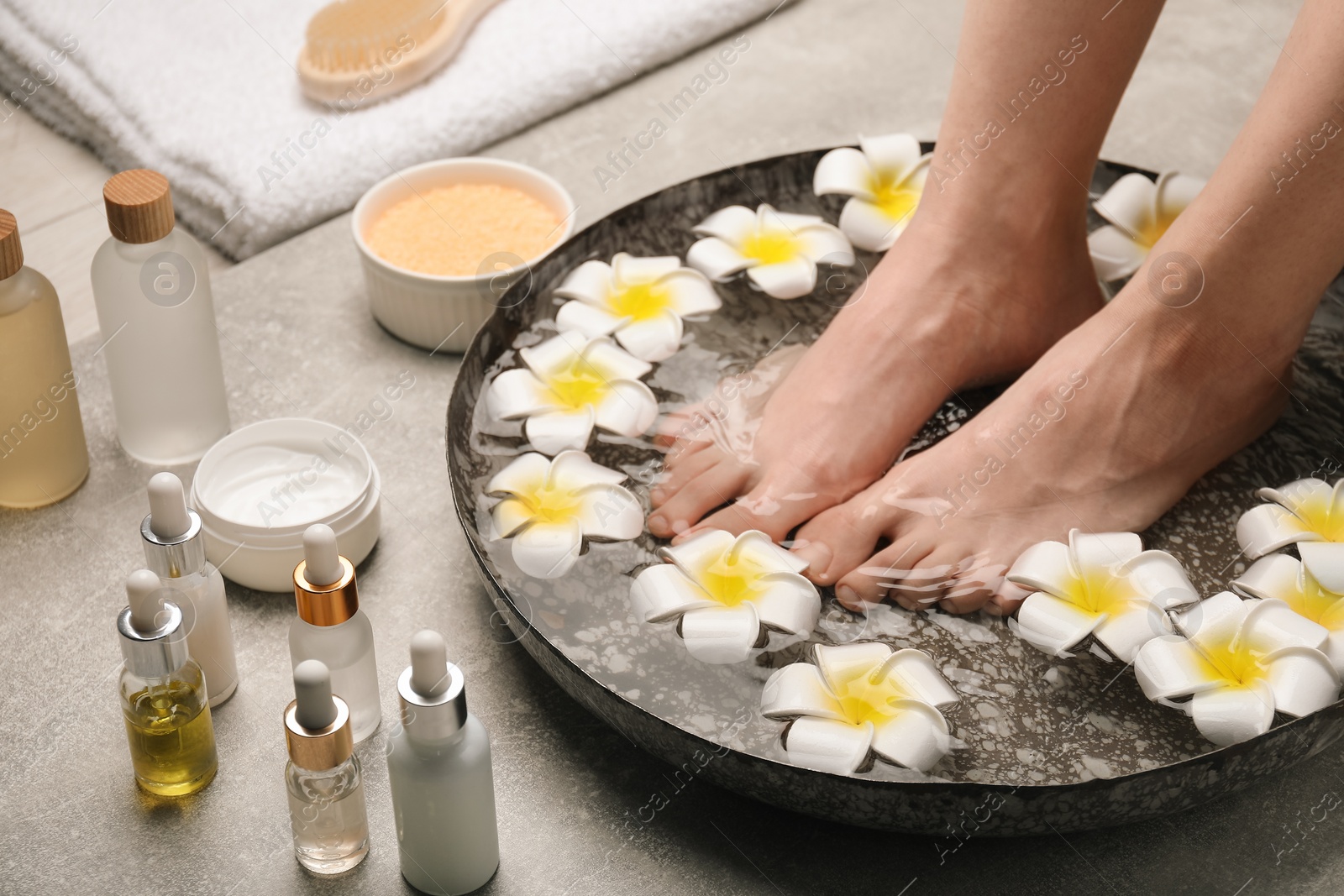 Photo of Woman soaking her feet in bowl with water and flowers on light grey floor, closeup. Spa treatment