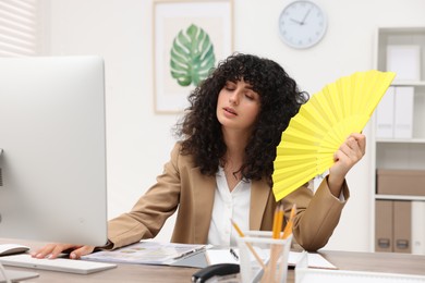 Photo of Young businesswoman waving hand fan to cool herself at table in office