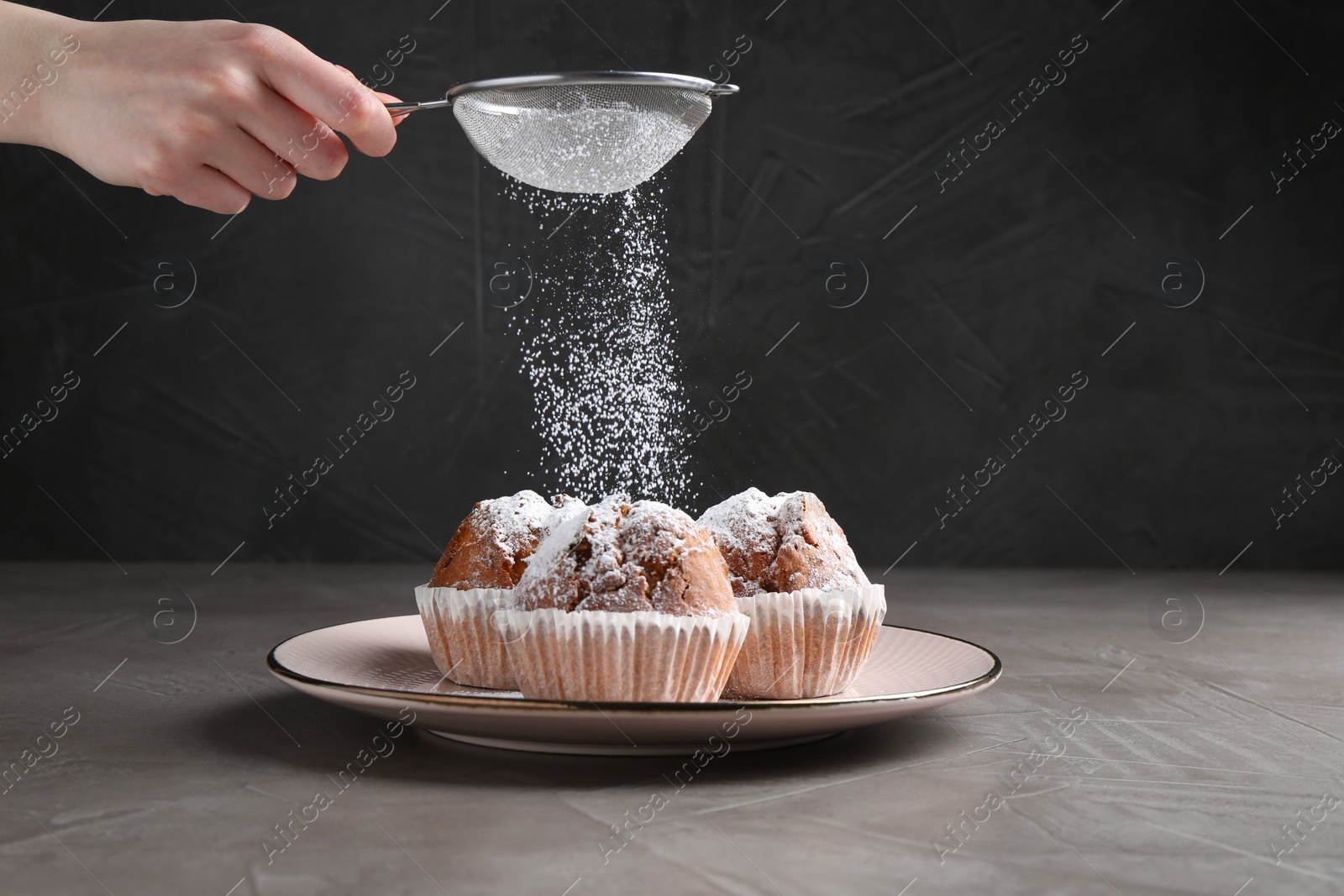 Photo of Woman with sieve sprinkling powdered sugar onto muffins at grey textured table, closeup