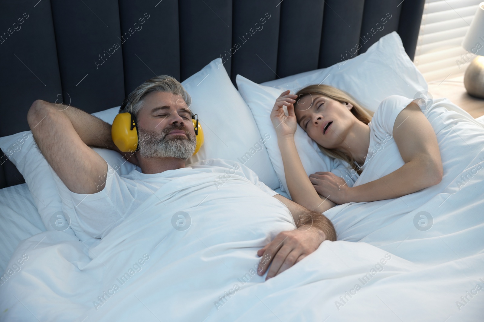 Photo of Smiling man with headphones lying near his snoring wife in bed at home