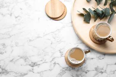 Mugs of coffee, stylish wooden cup coasters and eucalyptus branches on white marble table, flat lay. Space for text