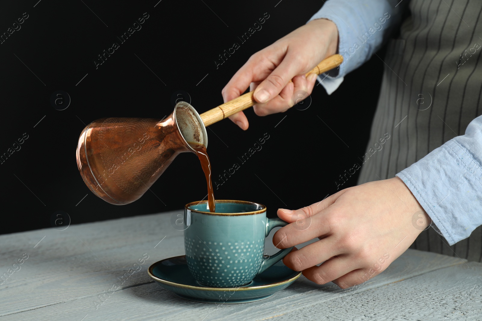 Photo of Turkish coffee. Woman pouring brewed beverage from cezve into cup at gray wooden table against black background, closeup