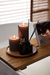 Photo of Wooden tray with burning candles, aroma diffuser and essential oil on bathtub in bathroom