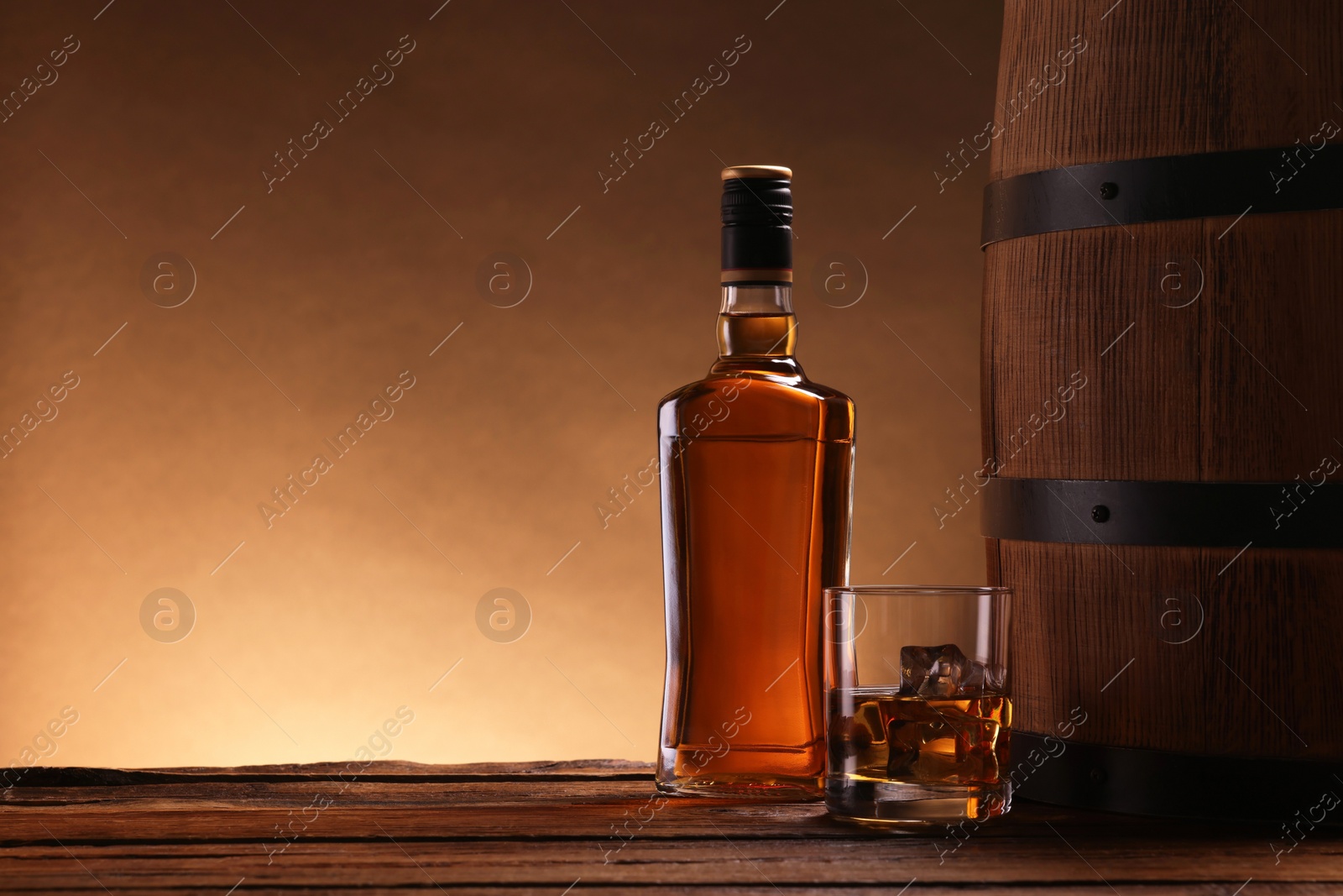 Photo of Whiskey with ice cubes in glass, bottle and barrel on wooden table, space for text