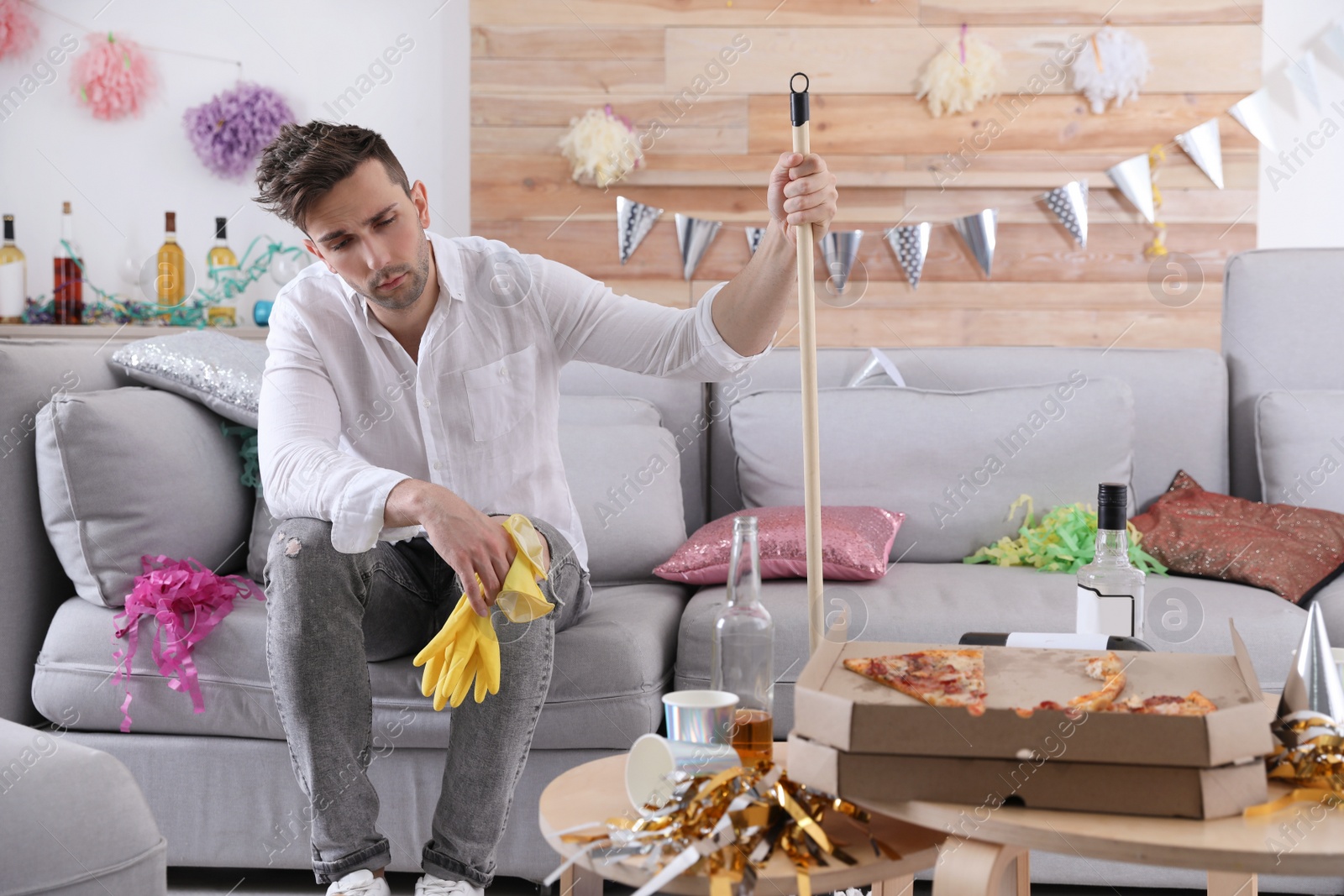 Photo of Young man with mop suffering from hangover in messy room after party