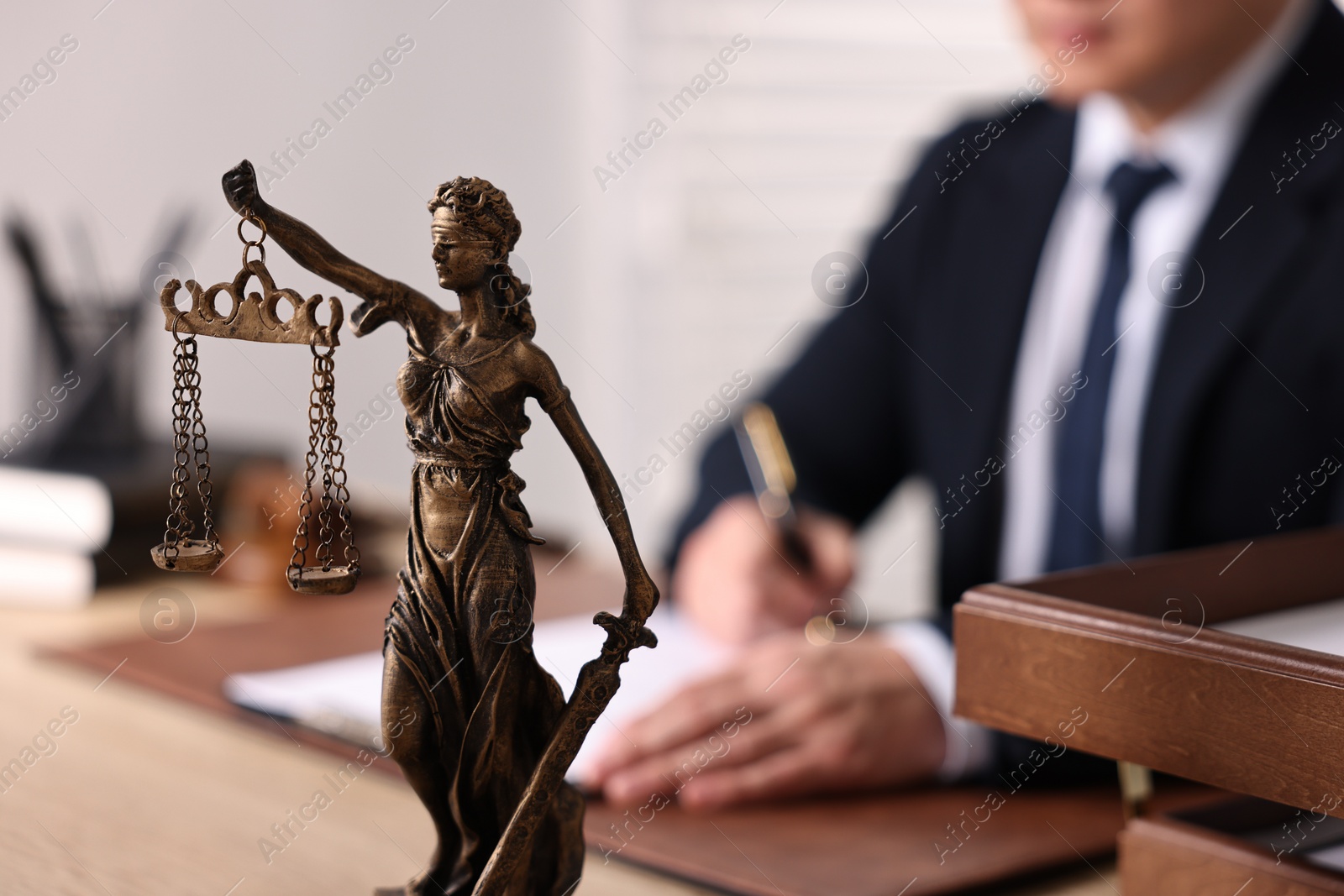 Photo of Notary writing notes at wooden table in office, focus on statue of Lady Justice