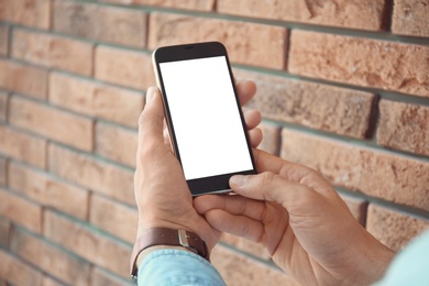 Photo of Young man holding mobile phone with blank screen in hands near brick wall