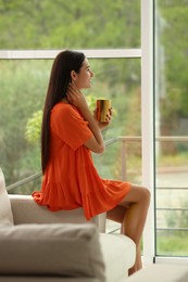 Photo of Young woman with cup of drink near sofa at home