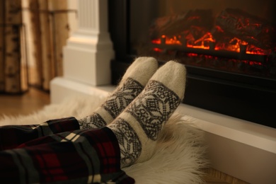 Woman in warm socks sitting near fireplace with burning wood at home, closeup