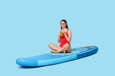 Happy woman with refreshing drink resting on SUP board against light blue background