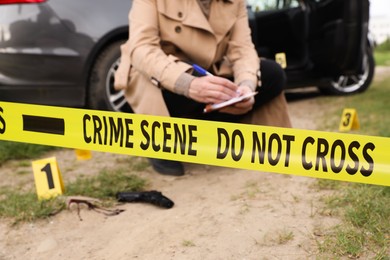 Photo of Professional detective examining crime scene outdoors, focus on yellow tape