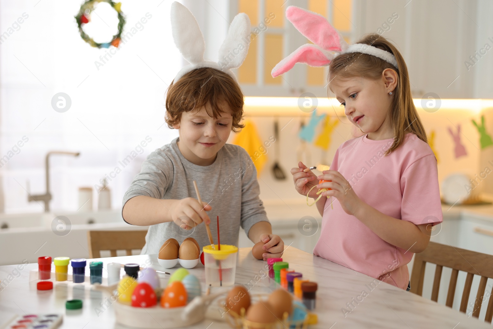 Photo of Easter celebration. Cute children with bunny ears painting eggs at white marble table in kitchen