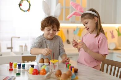 Photo of Easter celebration. Cute children with bunny ears painting eggs at white marble table in kitchen