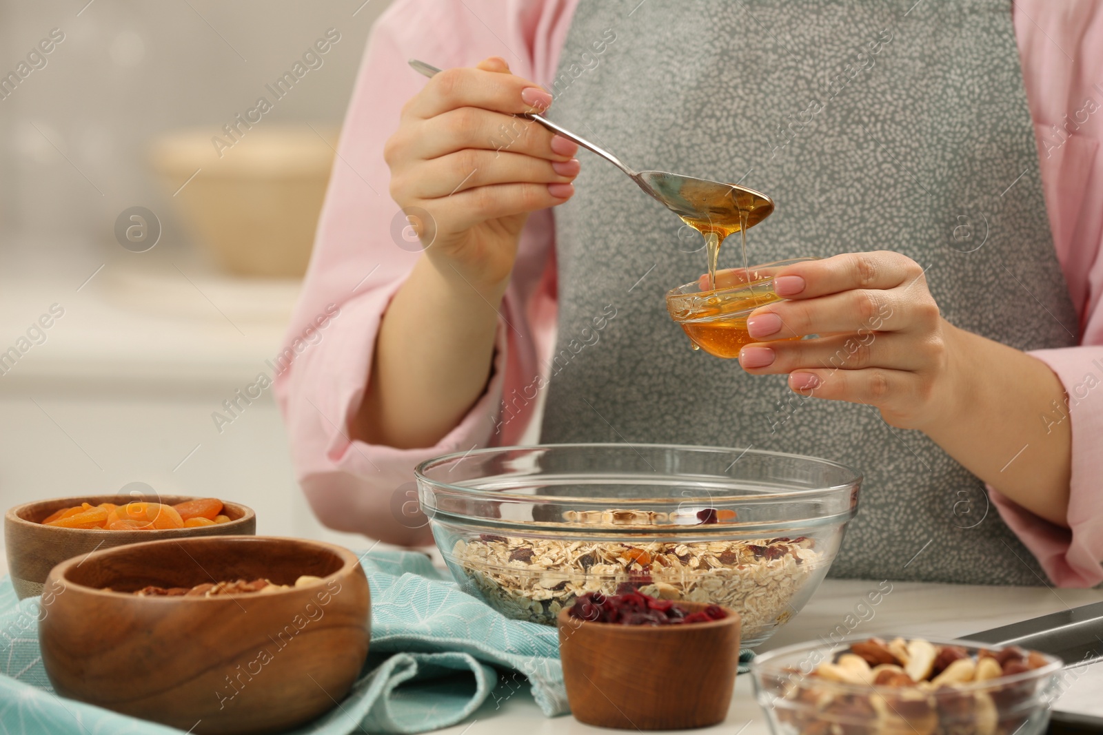 Photo of Making granola. Woman adding honey into bowl with mixture of oat flakes and other ingredients at table in kitchen, closeup