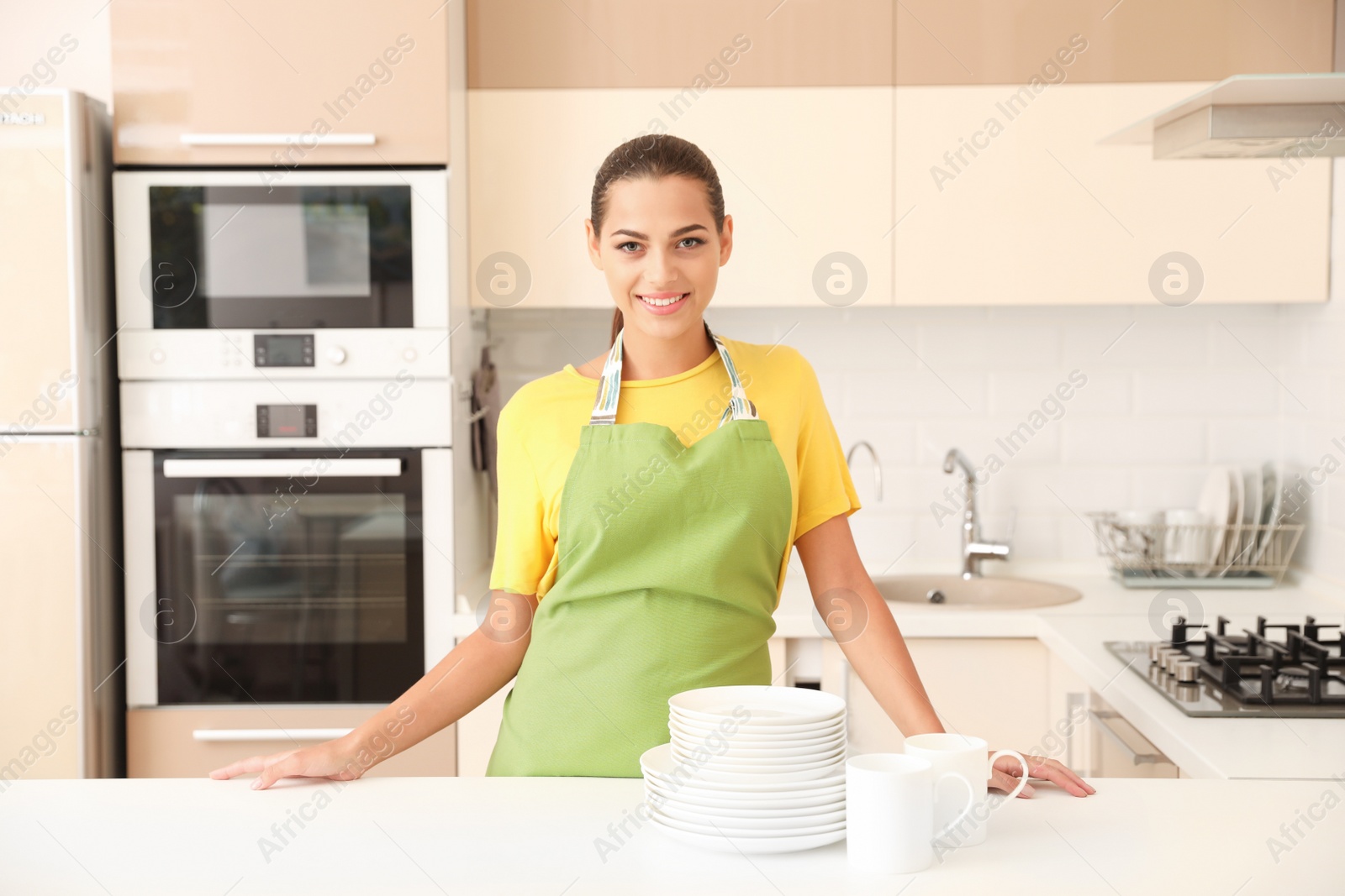 Photo of Beautiful young woman with clean dishes and cups at table in kitchen