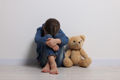 Photo of Child abuse. Upset girl with toy sitting on floor near white wall
