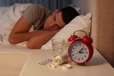 Photo of Man suffering from insomnia in bed at home, focus on pills and alarm clock