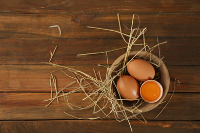 Raw chicken eggs and decorative straw on wooden table, top view