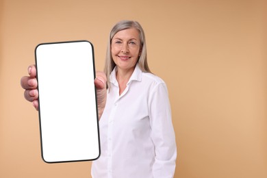 Happy mature woman showing mobile phone with blank screen on beige background. Mockup for design